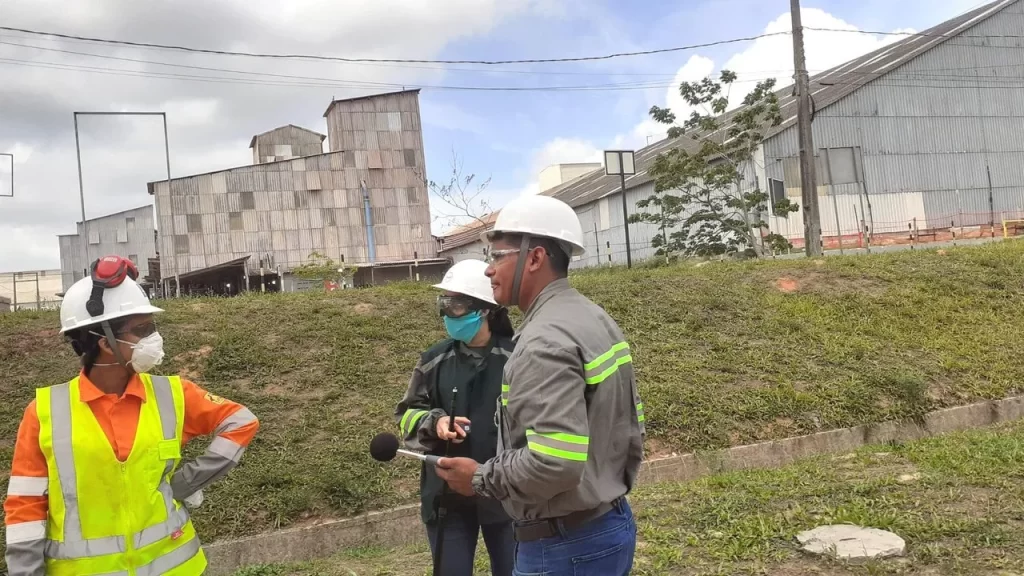 Três pessoas na área externa do empreendimento de fertilizante, paradas e utilizando EPI's - Equipamento de Proteção Individual mais ao fundo um armazém.