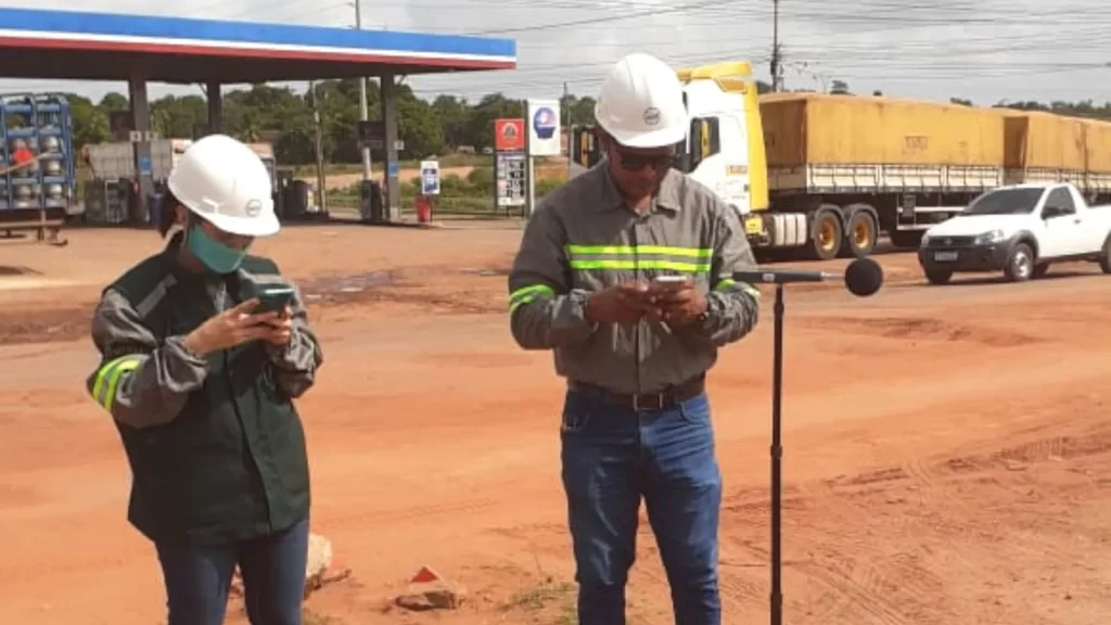 Duas pessoas formadas em engenharia em um segundo ponto fazendo o monitoramento de ruído ambiental na área exterior de uma empresa de fertilizante, mais atrás um posto d gasolina com um carro e caminhão.