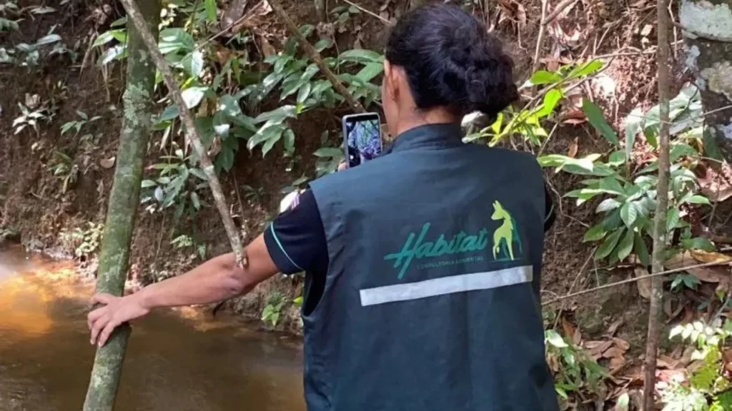 Mulher realizando no rio realizando teste de vazão em rio, fazendo registros fotográficos da área com celular na mão e colete.