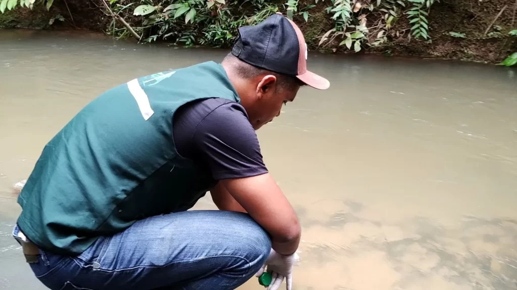 Engenheiro Ambiental agachado realizando procedimento de licenciamento coletando amostra de água do rio.