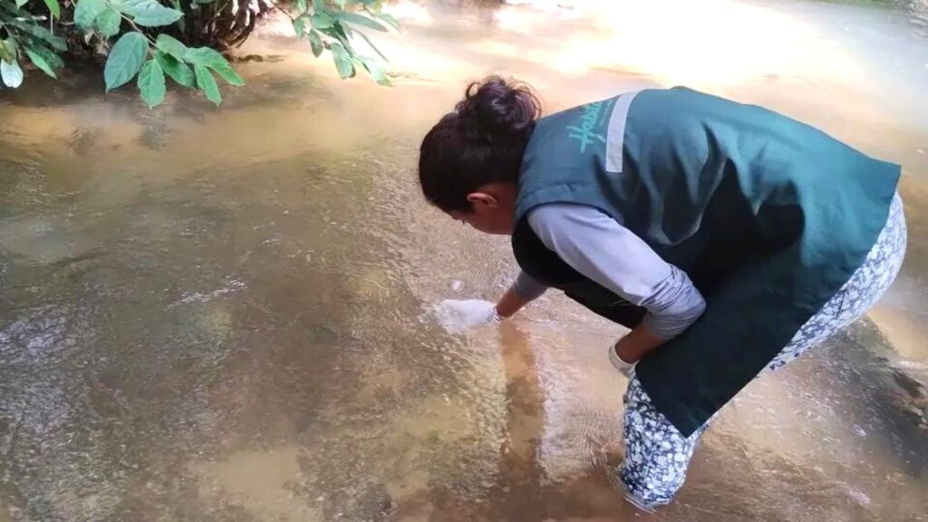 Geografa em rio realizando procedimento de coleta de recurso hídrico para análise próxima à vegetação.